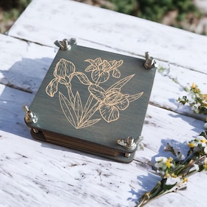 May include: A wooden flower press with a green stained finish and a gold iris flower design. The press is secured with four silver wing nuts. The press is sitting on a white wooden surface with some flowers.