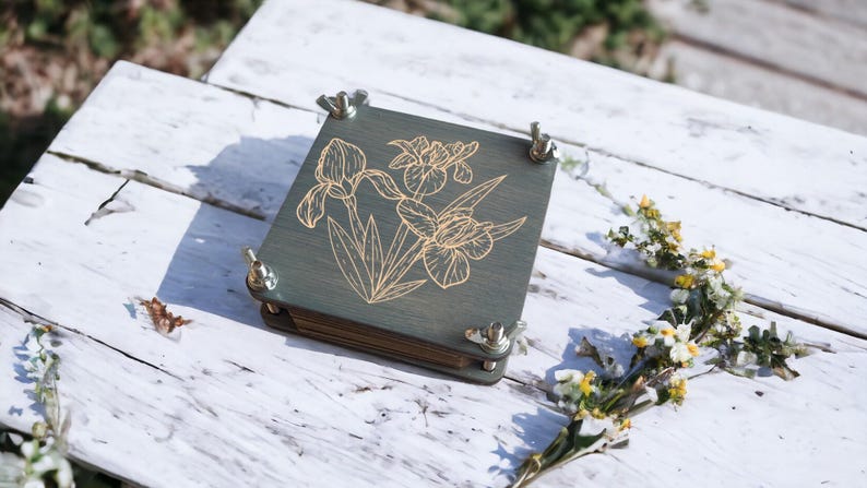 May include: A wooden flower press with a green stained finish and a gold iris flower design. The press is secured with four silver wing nuts. The press is sitting on a white wooden surface with some flowers.
