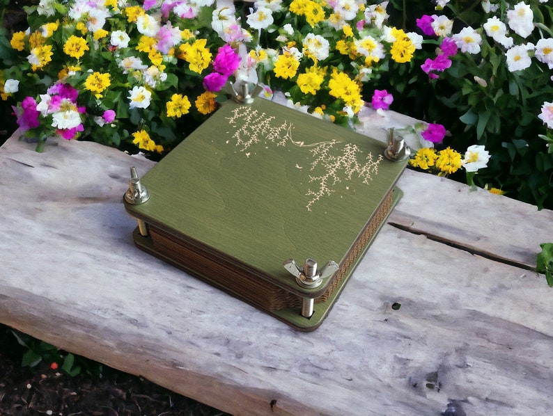May include: A green wooden flower press with a decorative gold design and metal hardware. The press is rectangular and has a stack of pages inside. The press is sitting on a wooden surface with colorful flowers in the background.