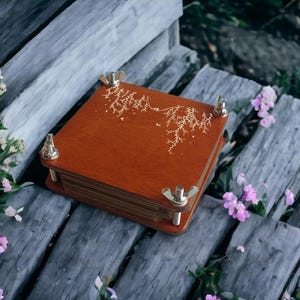 May include: A wooden plant press with brass hardware and a decorative gold design on the cover. The press is square and sits on weathered wooden planks, surrounded by greenery and small pink flowers. A tool for preserving botanical specimens.