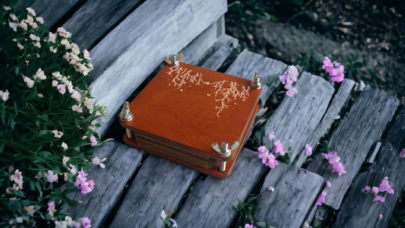 May include: A wooden plant press with brass hardware and a decorative gold design on the cover. The press is square and sits on weathered wooden planks, surrounded by greenery and small pink flowers. A tool for preserving botanical specimens.