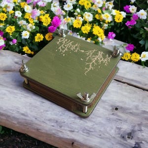 May include: A green wooden flower press with a decorative design on the top. The press is secured with four metal wingnuts. The press is sitting on a wooden surface with colorful flowers in the background.