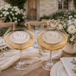 May include: Two champagne coupe glasses filled with a golden liquid, each with a circular label featuring the initials "A J" and the text "The Andersons 09.12.2026". The glasses are on a wooden table, with floral arrangements in the background.