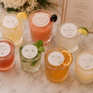 May include: An assortment of cocktails in crystal glasses, garnished with fruit and herbs. Each drink has a personalized circular label with initials or text. The drinks are arranged on a marble surface, with a decorative sign in the background.