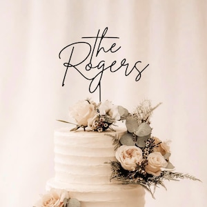 Peut inclure: Un gâteau blanc à plusieurs niveaux orné de fleurs et d'un cake topper noir avec l'inscription "the Rogers". Le gâteau est décoré de roses roses et blanches, de verdure et de feuilles d'eucalyptus.