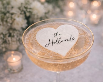May include: A champagne coupe filled with a bubbly, golden-hued beverage. A white heart-shaped drink topper with the text "the Hollands" floats on top. The glass has a gold rim. Soft lighting and flowers are in the background.