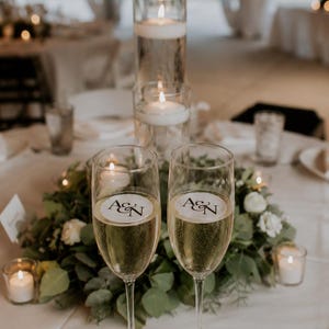 May include: Two champagne flutes filled with bubbly liquid, each with a circular sticker bearing the initials "A&N". The flutes sit on a white tablecloth, surrounded by greenery, white flowers, and lit candles, creating a celebratory atmosphere.