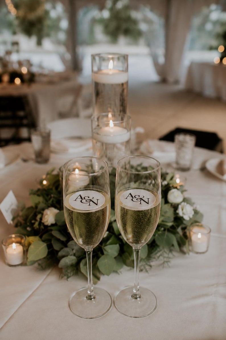 May include: Two champagne flutes filled with bubbly liquid, each with a circular sticker bearing the initials "A&N". The flutes sit on a white tablecloth, surrounded by greenery, white flowers, and lit candles, creating a celebratory atmosphere.