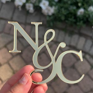 May include: A gold-coloured monogram featuring the letters "N & C" in a stylish script. The letters are held up against a blurred background of greenery and a brick surface. The monogram is likely for a wedding or special event.