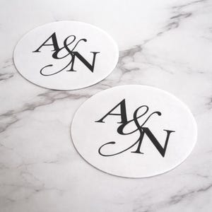 May include: Two white, oval coasters with the letters "A & N" in elegant black script. The coasters are on a marble surface, creating a sophisticated and personalized design.