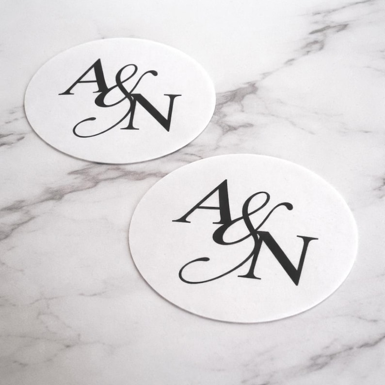 May include: Two white, oval coasters with the letters "A & N" in elegant black script. The coasters are on a marble surface, creating a sophisticated and personalized design.