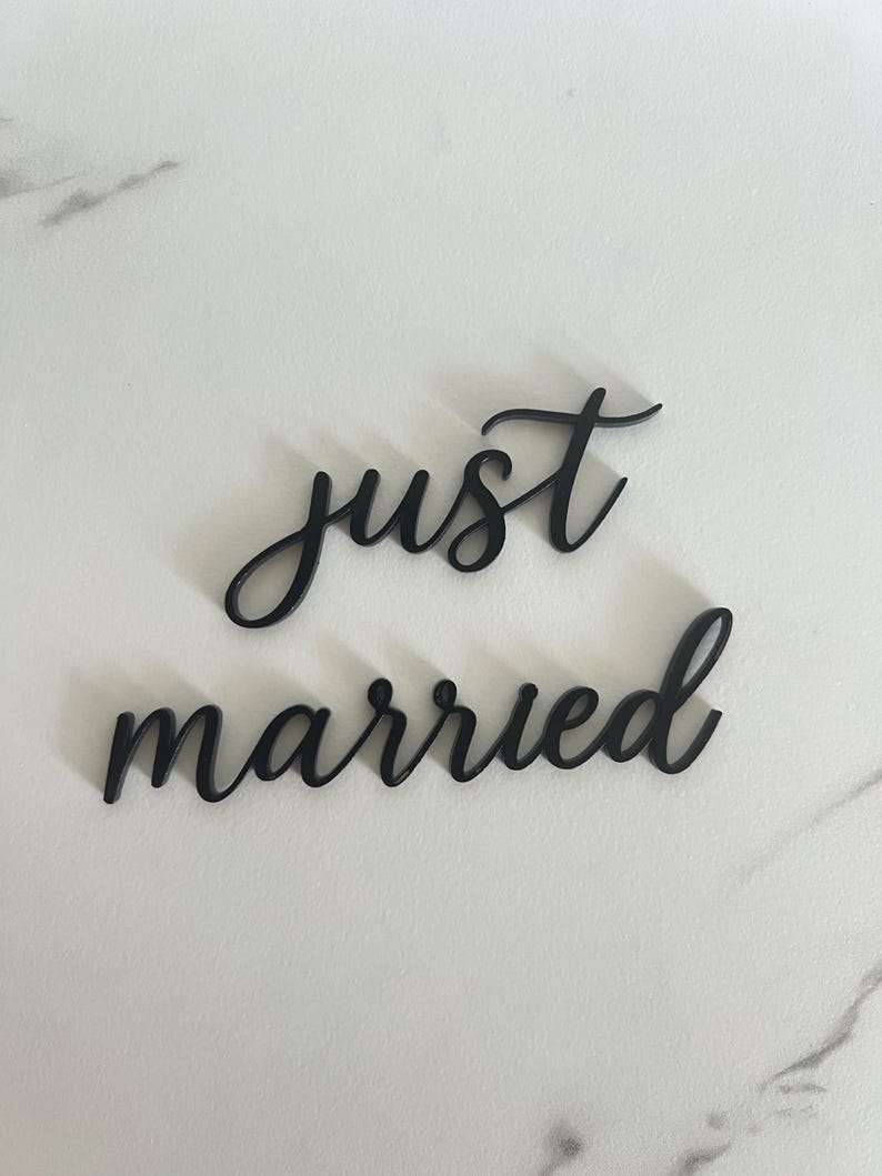 May include: Black cursive text that reads "just married" on a white marble surface. The words are separate and in a stylish font, perfect for wedding decor or photo props.