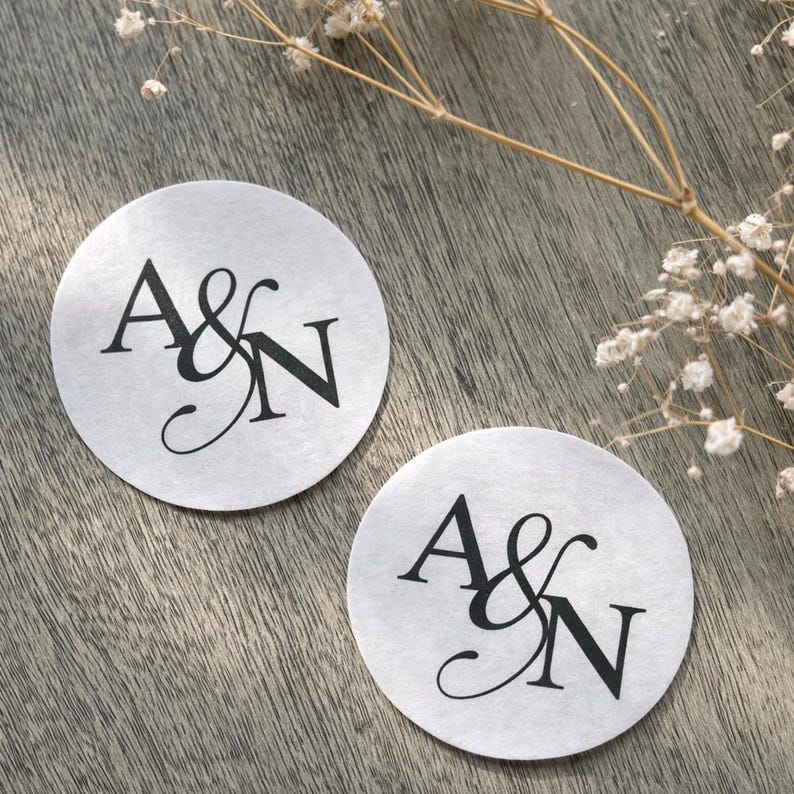 May include: Two round, light gray stickers with the letters "A&N" in black script. The stickers are on a wooden surface, with a sprig of dried flowers in the background. The stickers are likely for a wedding or other special event.