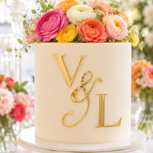 May include: A cylindrical cake with smooth off-white frosting, featuring large, reflective gold-colored letters V and L separated by an ampersand on the front. The top is decorated with a dense arrangement of fresh ranunculus flowers in pink, orange, yellow, and white.