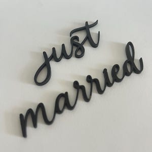 May include: Black cursive cut-out letters spelling "just" and "married" on a white surface. The letters are in a flowing script style, perfect for wedding decor or photo props. The words are separate.