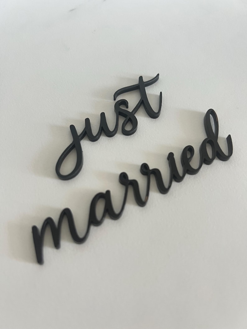 May include: Black cursive cut-out letters spelling "just" and "married" on a white surface. The letters are in a flowing script style, perfect for wedding decor or photo props. The words are separate.