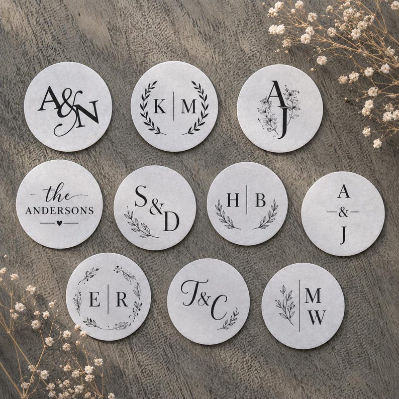 May include: A collection of personalized, round paper coasters with various initial and name designs in black ink. Some coasters feature floral or laurel wreath accents, while others display initials or names like "The Andersons."