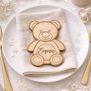 May include: A wooden teddy bear-shaped place card with the name "Emma" engraved on it, resting on a white napkin atop a white plate. The table setting includes gold cutlery, a cupcake, and a heart-shaped cookie.