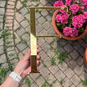 May include: A gold-coloured letter "F" is held up against a backdrop of brick paving and greenery. The letter is reflective, and the gold colour gives it a luxurious appearance. A pot of pink flowers is in the background.