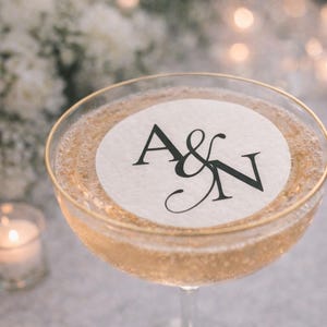 May include: A champagne coupe filled with a sparkling beverage, featuring a gold rim and a white circular label with the black letters "A&N". The background includes soft white flowers and blurred candles.