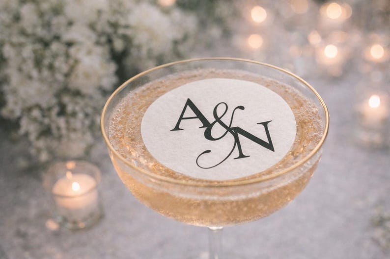 May include: A champagne coupe filled with a sparkling beverage, featuring a gold rim and a white circular label with the black letters "A&N". The background includes soft white flowers and blurred candles.