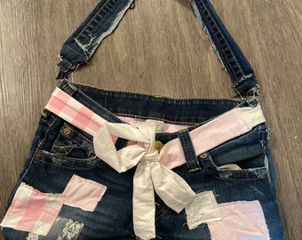 May include: A denim handbag with a pink and white checked strap and a white bow. The bag is made from repurposed denim and has patches of pink and white fabric.