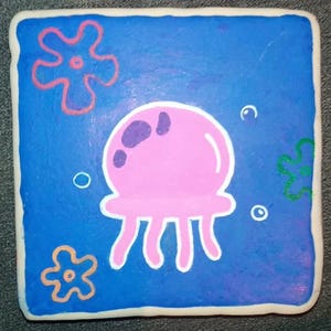 May include: A hand-painted square tile with a blue background featuring a pink jellyfish with purple spots and white tentacles. The tile also has three orange and pink flowers and a green seaweed.