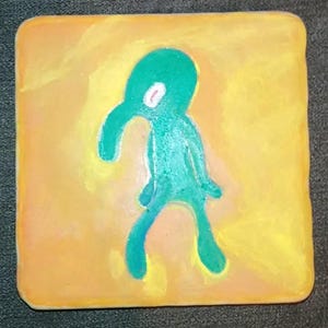May include: A hand-painted square tile with a yellow background and a green cartoon character with a long nose and one eye. The character is standing on the tile with its arms outstretched.