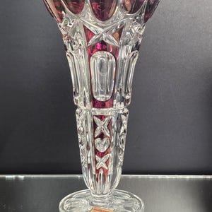 May include: A clear crystal vase with a flared top and a round base. The vase features a ruby red interior and accents, with a heart-shaped rim and intricate cut glass designs. The vase is approximately 10 inches tall.