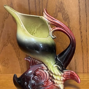 May include: A ceramic pitcher shaped like a fish with a green, black, and red glaze. The pitcher has a spout and a handle.