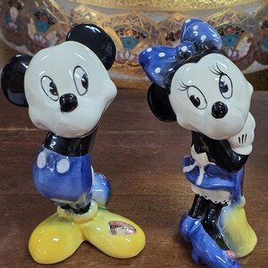 May include: A pair of ceramic Mickey and Minnie Mouse figurines. Mickey is in blue shorts and yellow shoes. Minnie wears a blue polka dot bow and dress. Both have black ears and are smiling.