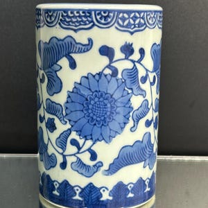 May include: A cylindrical ceramic vessel featuring a white base with a blue hand-painted floral pattern. The design includes large central chrysanthemum-like flowers surrounded by scrolling leaves and vines, with decorative borders at the top and bottom.