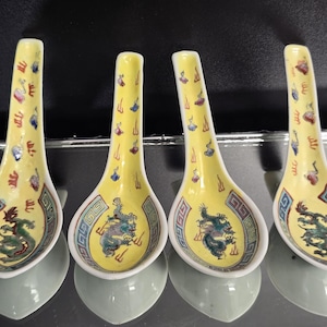May include: Four ceramic soup spoons with a yellow background, each featuring a green dragon motif in the bowl and a geometric blue and green border. The handles are decorated with small, colorful scattered symbols and flame-like patterns.