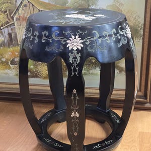 May include: Black, round stool with floral inlay. The top and sides feature intricate white and pale green floral designs. The stool has a sturdy, curved leg design and is likely made of wood.