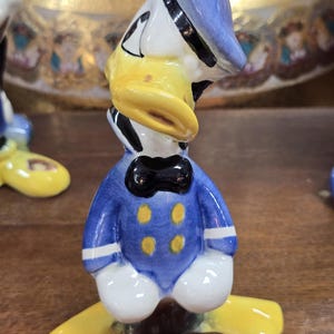 May include: A ceramic figurine of Donald Duck, featuring a blue sailor suit with yellow buttons, a black bow tie, and a blue hat. The duck has a yellow beak and feet, with white and black accents. The figurine is a collectible item.