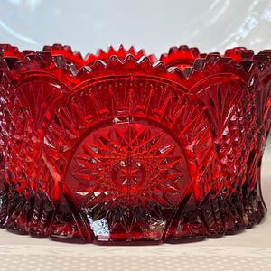 Puede incluir: Un cuenco de cristal rojo vibrante con un patrón prensado detallado, un motivo central de estrella, facetas geométricas en forma de diamante y un borde dentado. El cuenco tiene una base circular y una construcción gruesa y translúcida.