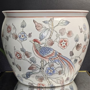 May include: A white ceramic planter with a hand-painted design featuring a bird perched on a branch with flowers. The bird has red, blue, and pink feathers. The planter has a red rim.