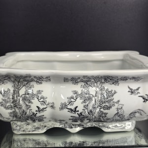 May include: A rectangular white ceramic planter with scalloped edges and a black toile-style print featuring detailed illustrations of trees, birds, and roosters. The vessel has a wide opening and a raised base with a matching black and white pattern.