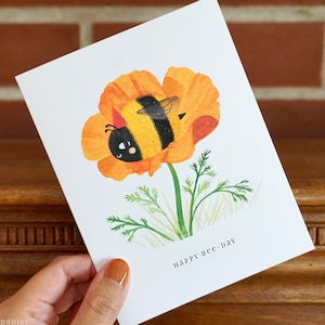 May include: A greeting card with a watercolor illustration of a bee inside a yellow flower. The text "happy bee-day" is printed on the card.