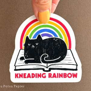 May include: A black cat kneading on a book with a rainbow arch above it. The text "Kneading Rainbow" is printed below the cat. The sticker is white with a red border.