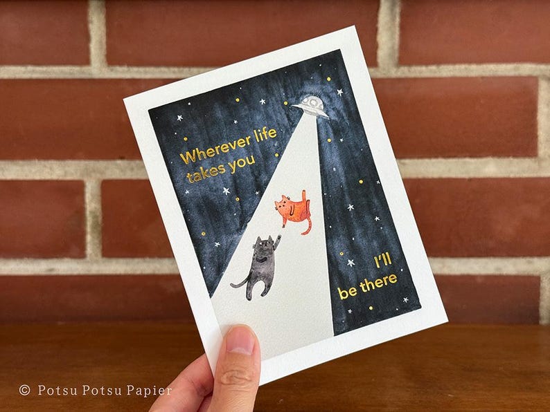 Close up of greeting card of hand painted illustration of two cats sitting on a fence and looking up at the moon. Message and embellishments in gold pressed-foil.