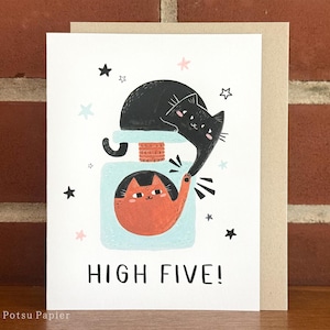 May include: A greeting card with a black cat and an orange cat giving each other a high five. The card has a blue background with white stars and the text "HIGH FIVE!" in black.