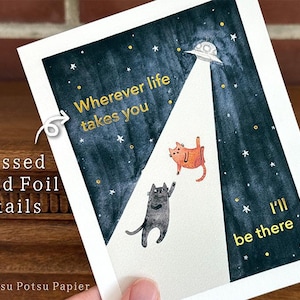 May include: An art print with a dark blue night sky, a UFO, and two cats. Gold foil text reads "Wherever life takes you, I'll be there". The print has gold foil details. The print is on white paper.