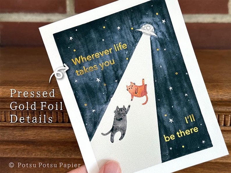 May include: An art print with a dark blue night sky, a UFO, and two cats. Gold foil text reads "Wherever life takes you, I'll be there". The print has gold foil details. The print is on white paper.