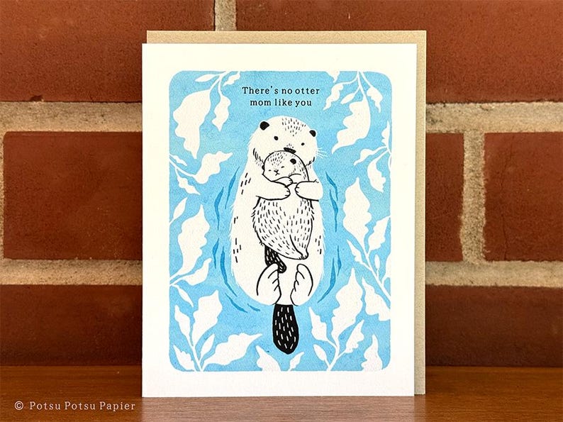 Hand illustrated card of an otter holding onto its cub while floating through a blue sea of seaweed. 

Message: There's no otter mom like you

Includes a2 kraft envelope.