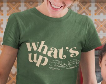 Peut inclure: Une femme et une jeune fille cuisinent ensemble dans une cuisine. La femme porte un t-shirt vert avec le texte "what's up" dans une police rétro. La fille porte un t-shirt blanc avec un chat de dessin animé dessus. Elles sourient toutes les deux et s'amusent.