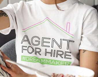 Könnte beinhalten: Eine Frau mit Brille trägt ein weißes T-Shirt mit dem Text "AGENT FOR HIRE #DREAMMAKER". Sie sitzt auf einem Sofa und telefoniert.