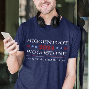 Op de afbeelding: Een marineblauw t-shirt met de tekst "Higgentoot 2024 Woodstone Anyone But Hamilton" gedrukt in wit, rood en blauw.