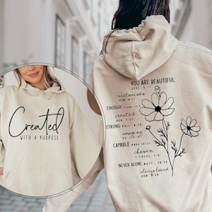 May include: Sand-colored hoodie with the text "Created With A Purpose" on the front. The back features a floral design with the words "You Are Beautiful," and other inspirational words and Bible verses. A comfortable, casual garment.