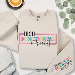 May include: A beige sweatshirt with the text "HIGH FUNCTIONING introvert" in colorful letters. The sweatshirt has a folded pair of blue jeans on top of it and a cup of coffee in the background.